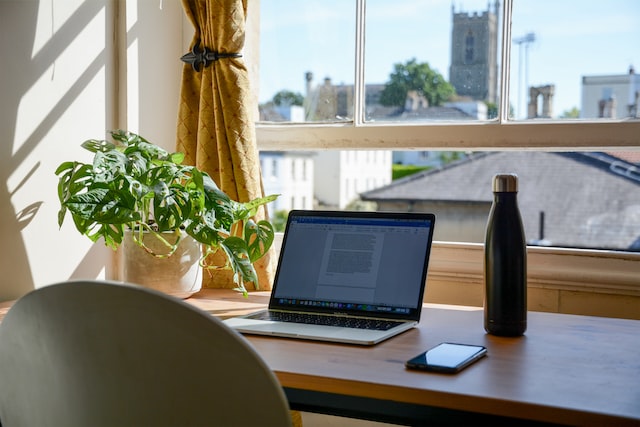Desk set up in front of a window with an open laptop, water bottle, and iphone